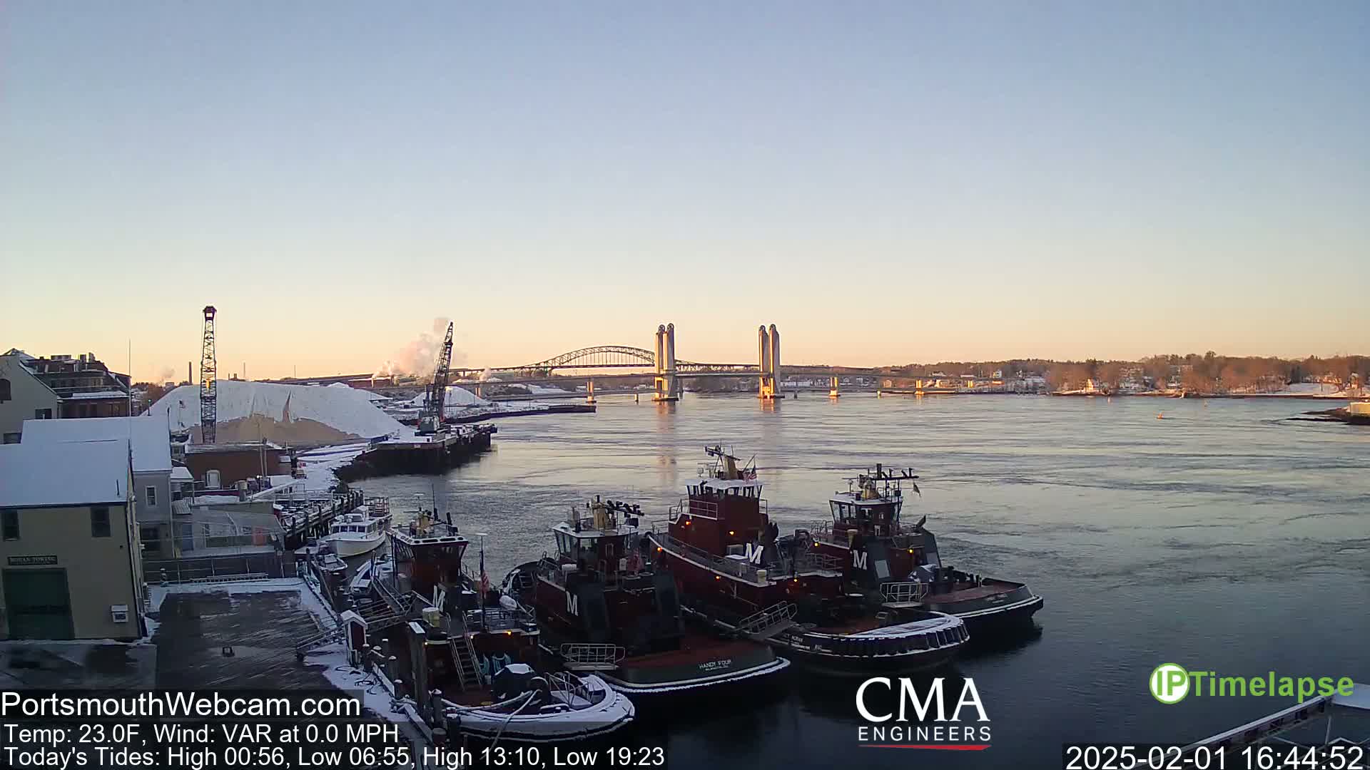 Several tugboats are moored at a snow-covered dock, with a bridge and a snow-covered hill in the background under a clear, twilight sky.