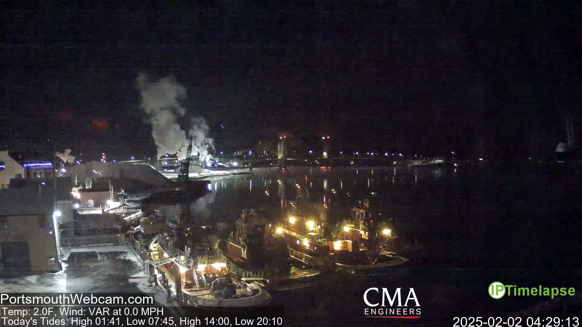 Several tugboats are moored at a dock at night, with smoke rising from industrial structures in the background under a clear, dark sky.