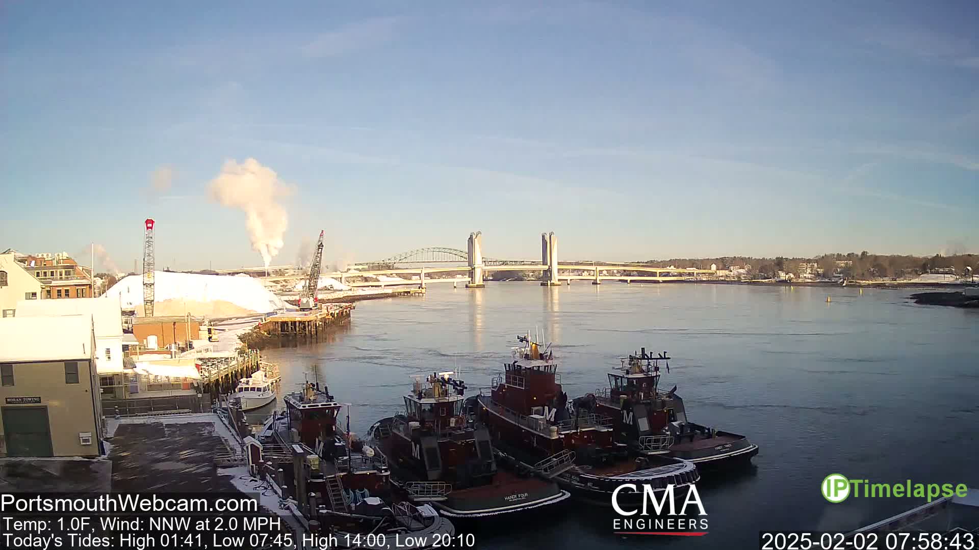 Several tugboats are docked at a waterfront, with a large pile of white material and a bridge in the background on a clear, cold day.