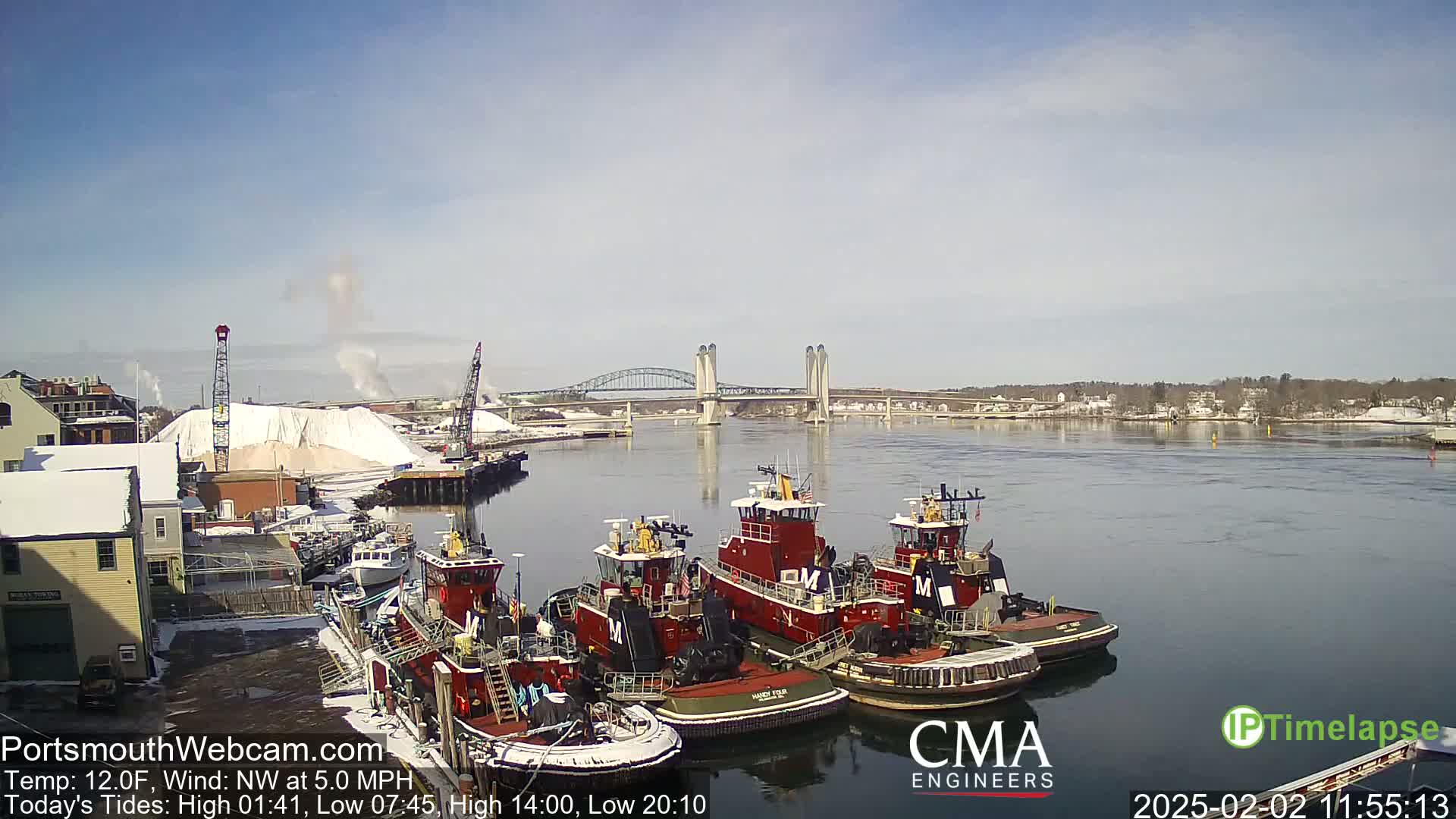Several tugboats are moored at a dock next to a snow-covered waterfront, with a bridge spanning a river in the background under a mostly clear sky.