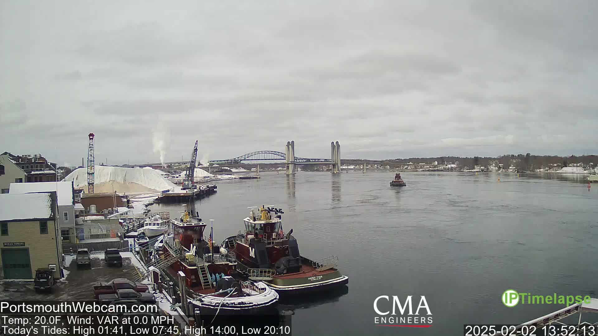 A wide shot of a harbor with two tugboats docked, a large pile of white material, and a bridge in the background under an overcast sky.