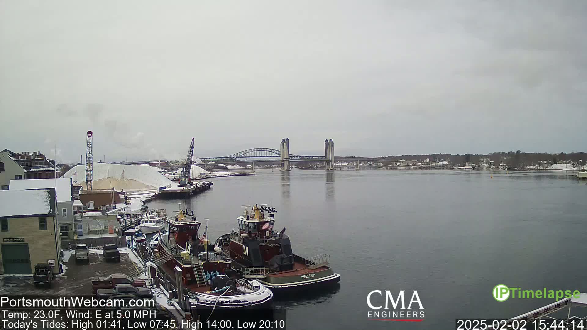 Two tugboats are docked near a snow-covered industrial area, with a bridge spanning a calm river under an overcast sky.