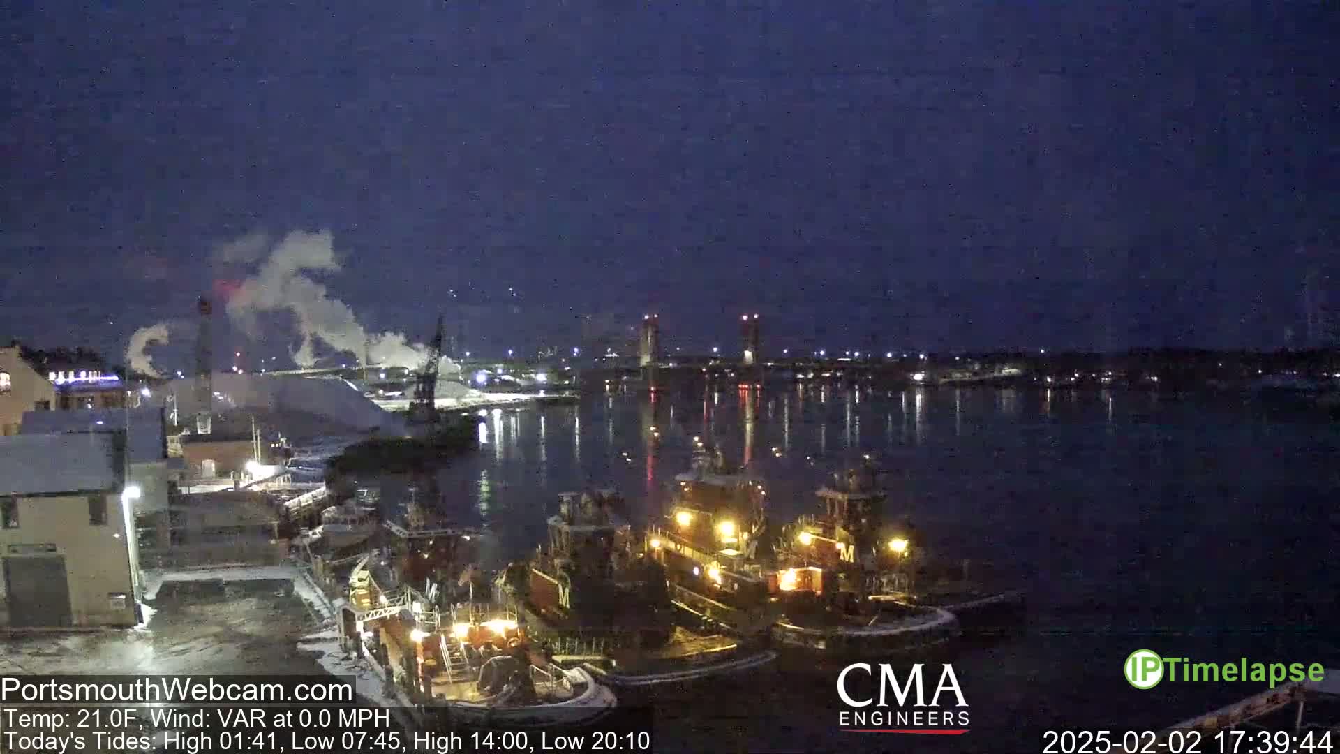 Several tugboats are moored at a dock at night under a dark sky, with smoke rising from industrial structures in the background.