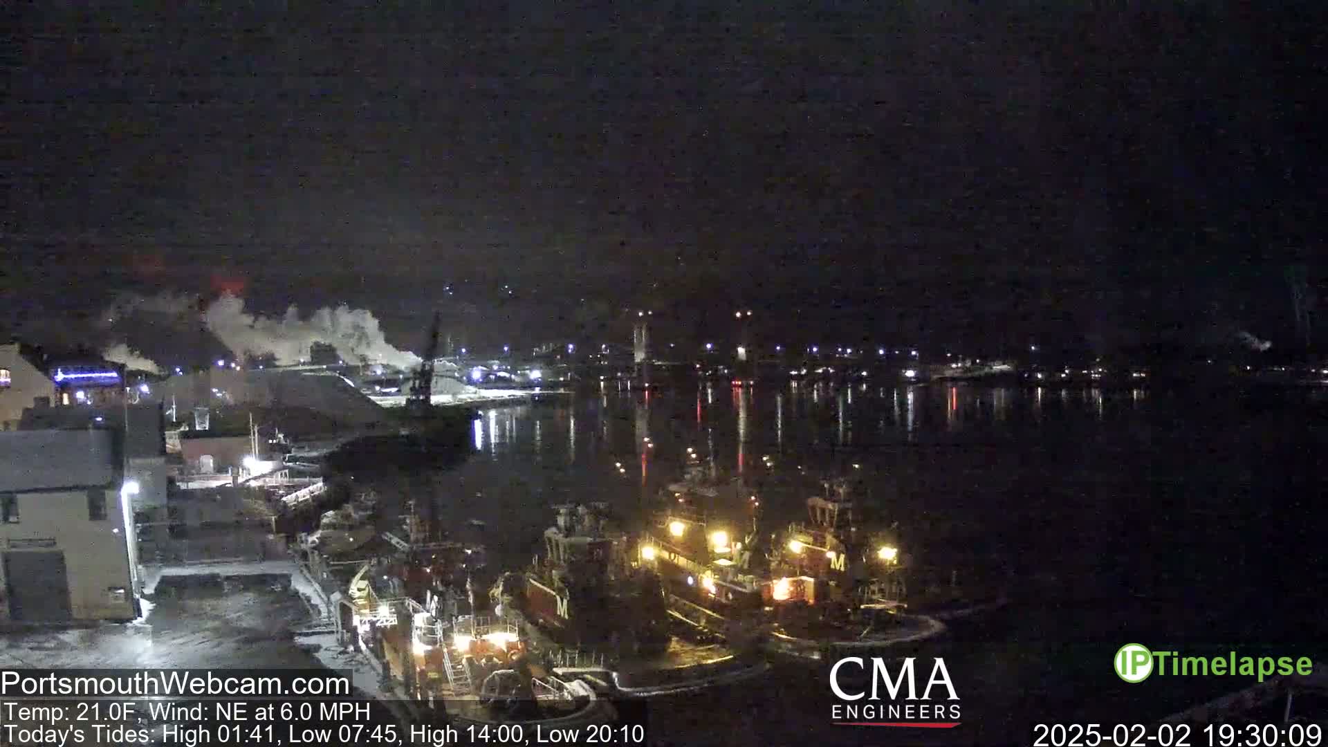 A nighttime harbor scene shows several tugboats moored near a snow-covered dock, with industrial lights and smoke in the distance under a dark, clear sky.