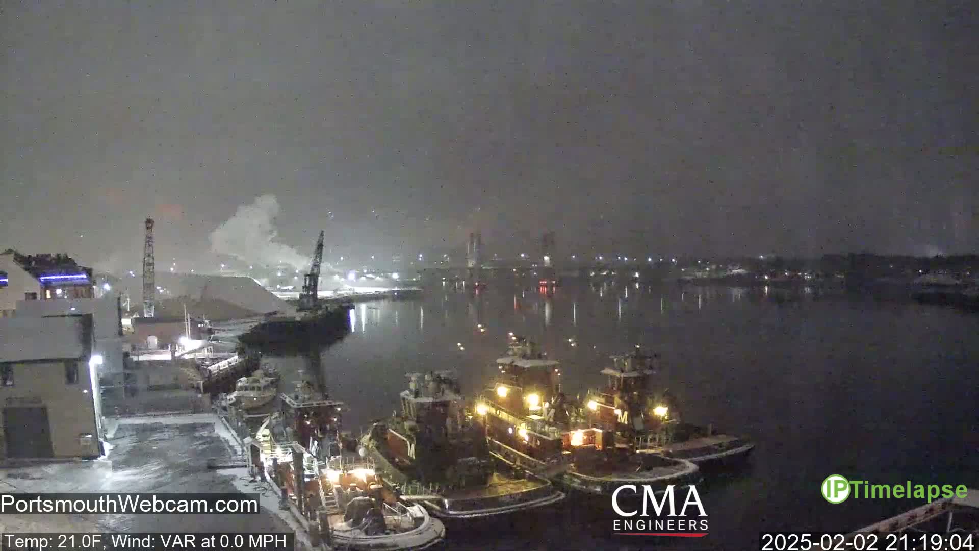 Several tugboats are docked at a snow-covered harbor at night during a light snowfall.