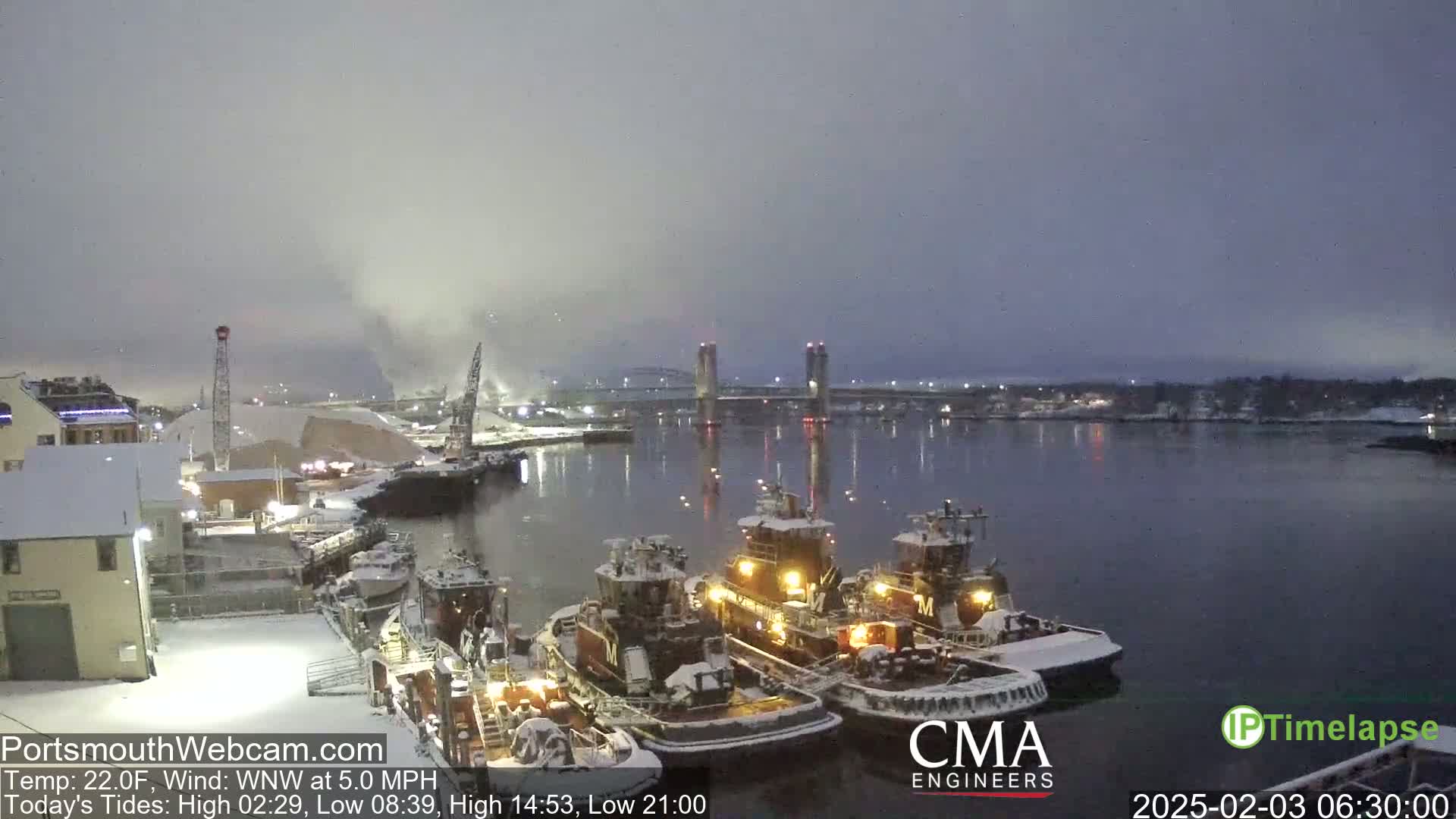 A snow-covered harbor scene at twilight shows several tugboats moored near a pier, with a bridge in the distance.
