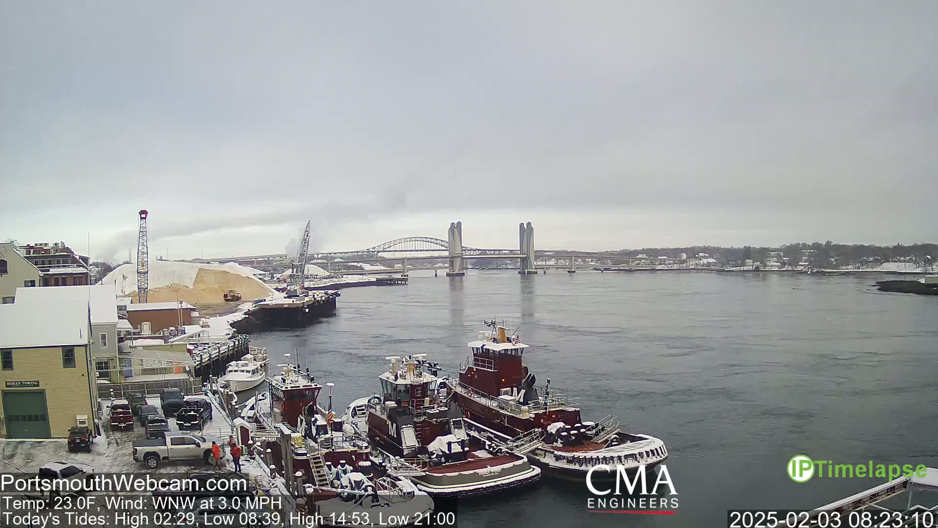 A snowy waterfront scene shows several tugboats docked near a large pile of sand, with a bridge spanning a river in the background under an overcast sky.