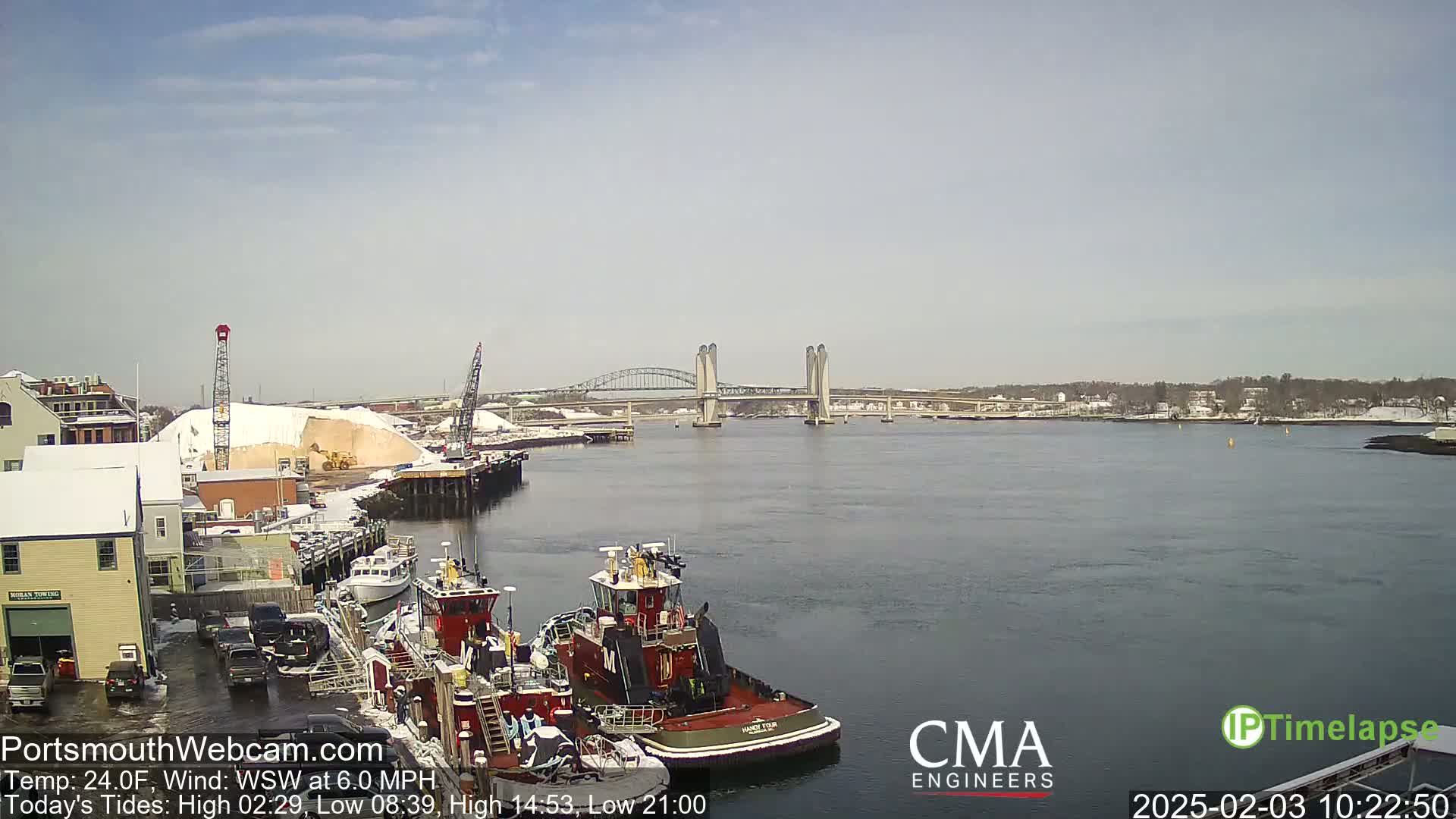 A harbor scene on a partly sunny day shows several tugboats docked near a snow-covered industrial area with a large bridge in the distance.