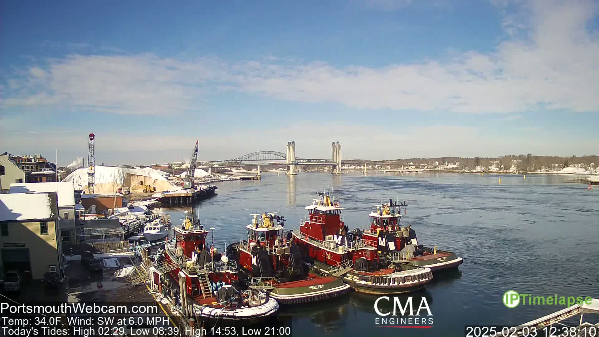 Several tugboats are moored at a dock near a bridge on a sunny day with a light southwest wind.