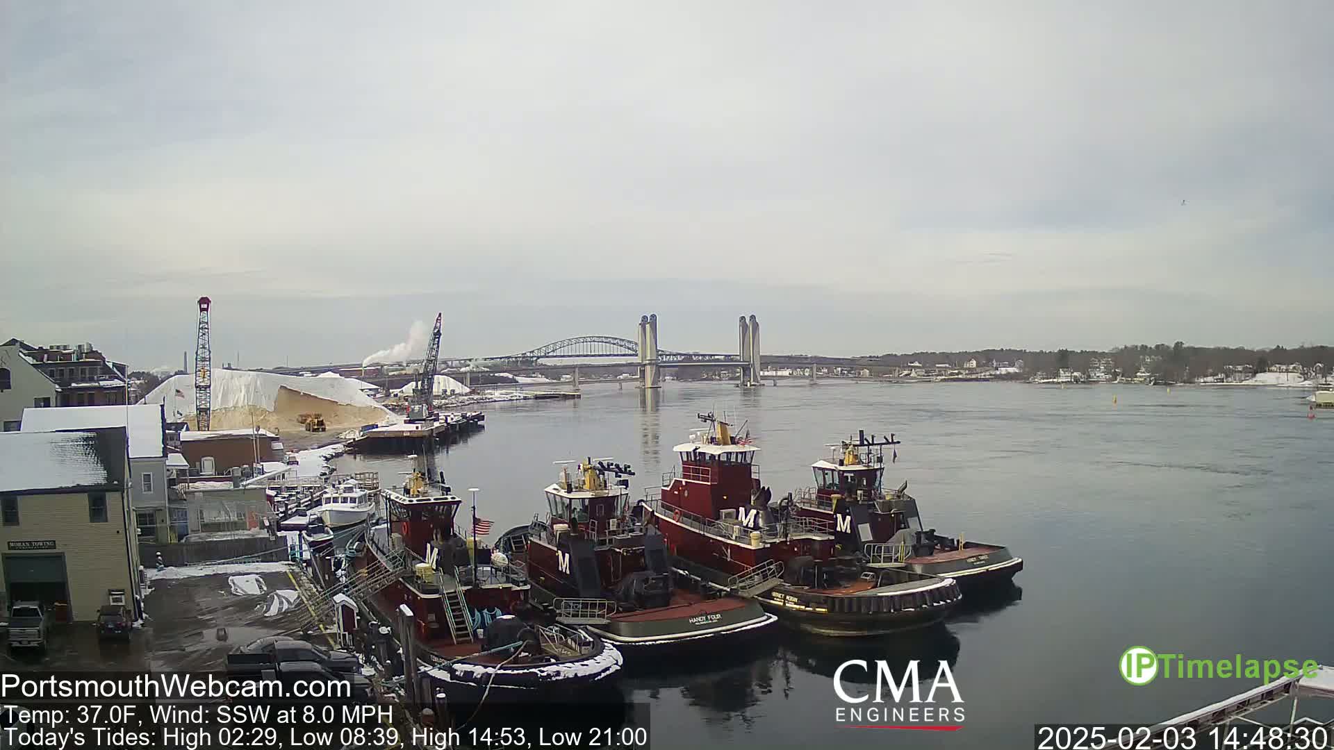 Several tugboats are moored at a dock near a bridge, with a pile of what appears to be salt in the background under an overcast sky.
