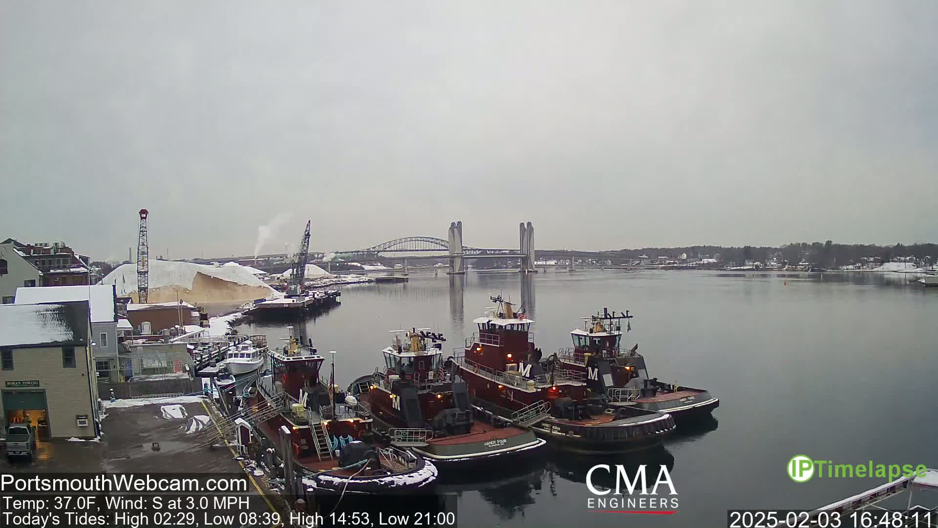Three tugboats are moored at a dock in a harbor under an overcast sky, with a bridge in the distance and snow on the ground.