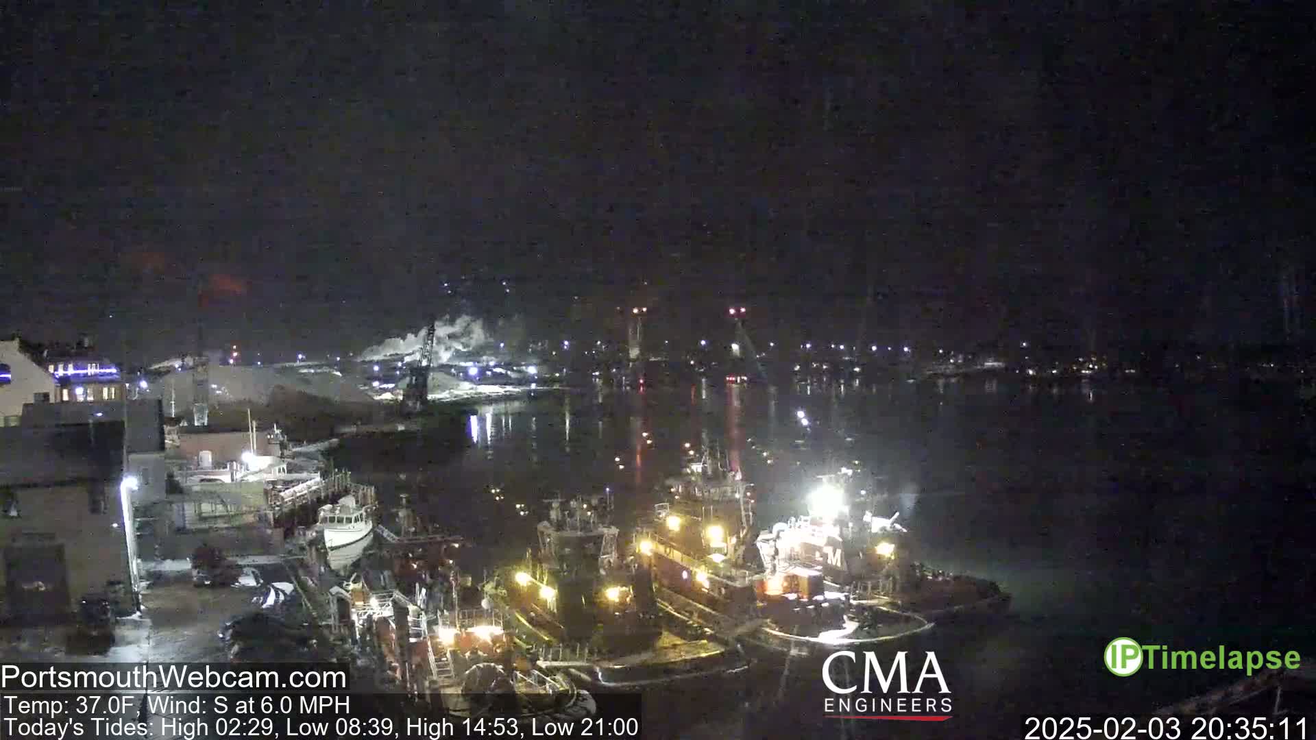 A nighttime harbor scene showing several tugboats moored near a waterfront, under a dark, clear sky.