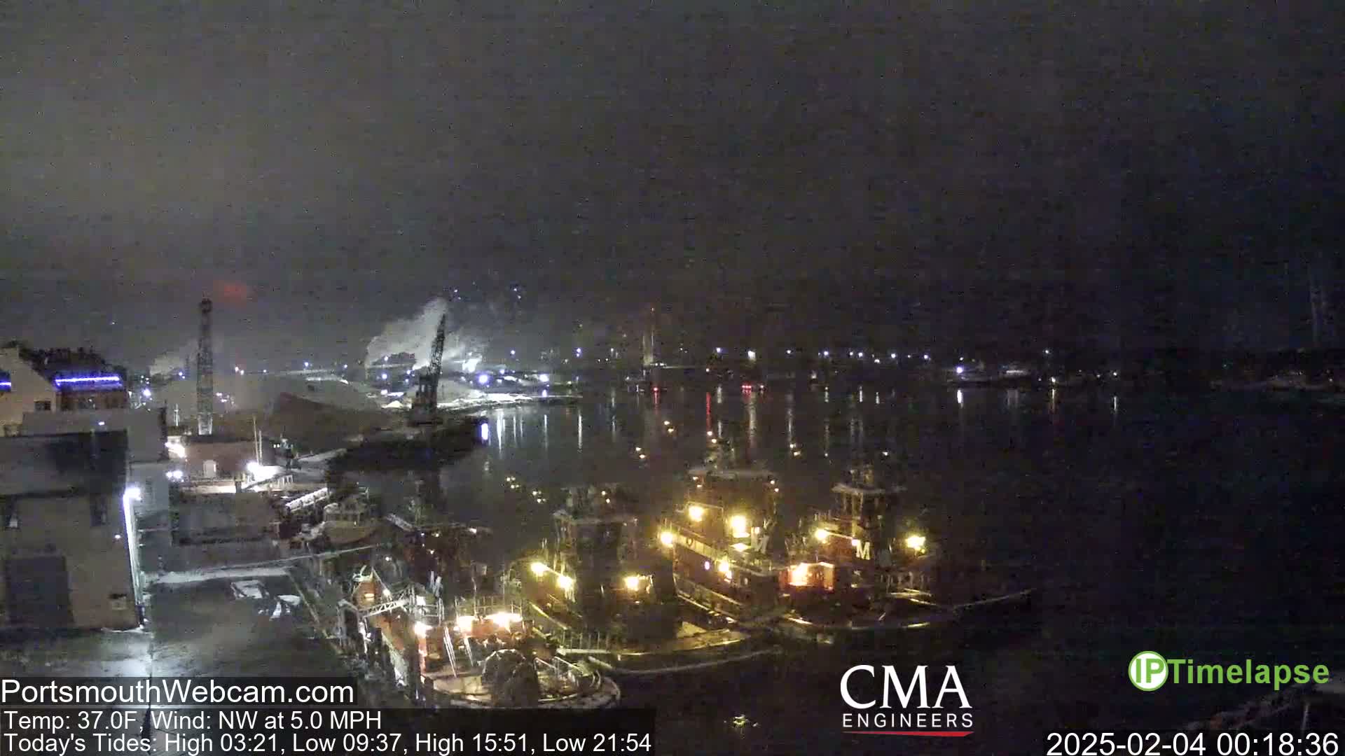 A nighttime harbor scene shows several tugboats docked near a waterfront industrial area under a dark, overcast sky.