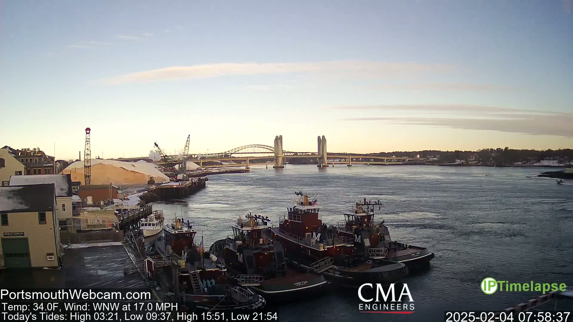 Several tugboats are moored at a dock near a bridge and a large pile of what appears to be snow or salt under a partly cloudy sky with a light wind.