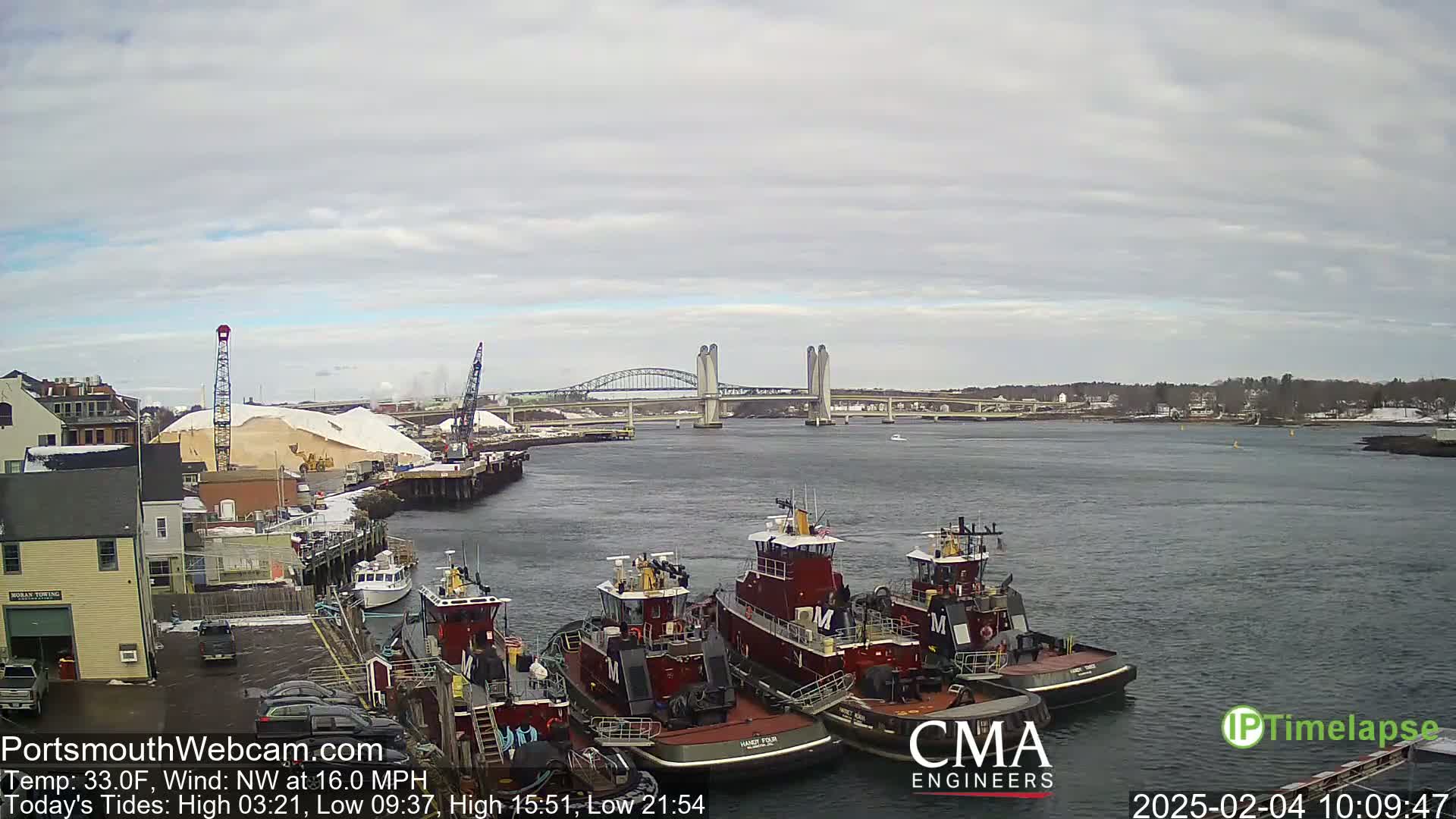 A harbor scene under a cloudy sky shows several tugboats moored at a dock, with a large bridge and a pile of snow visible in the background.