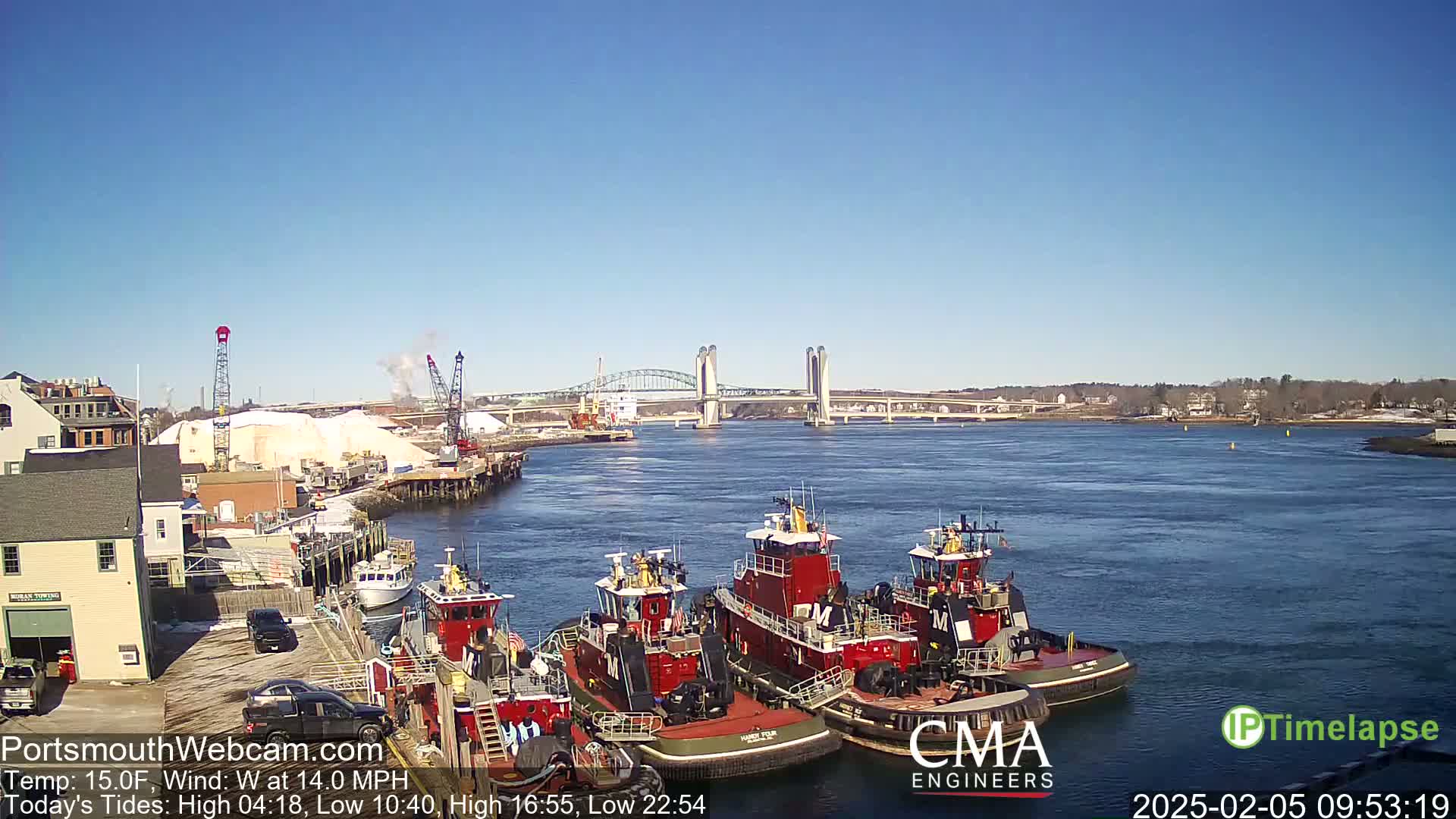 Several tugboats are docked at a waterfront, with a large bridge spanning a river under a clear blue sky.