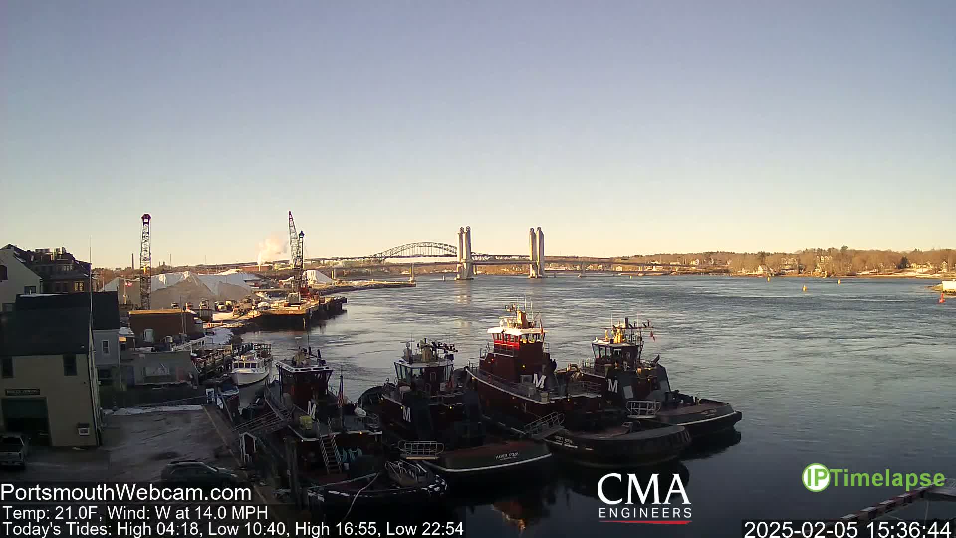 Several tugboats are moored at a dock next to a river, with a bridge and distant shoreline visible under a clear sky.