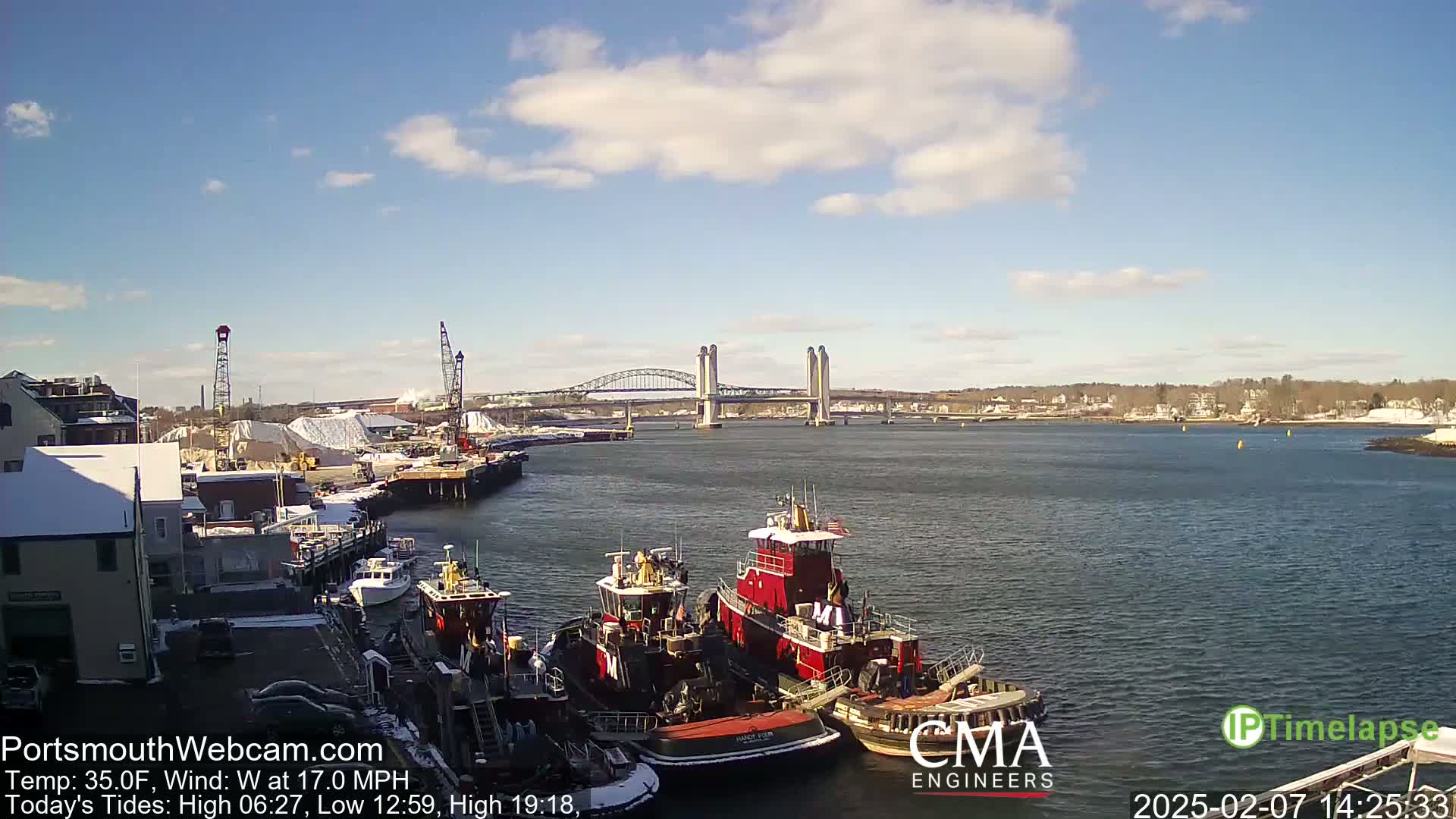 Several tugboats are moored at a dock next to a body of water under a partly cloudy sky, with a double-lift bridge visible in the distance.