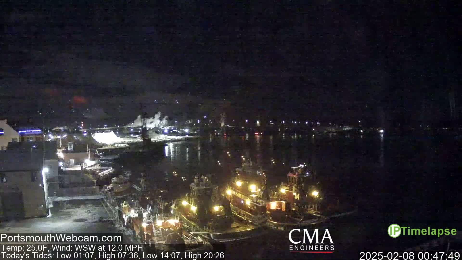 At night, several tugboats are moored at a dock, with a calm body of water and a distant, dimly lit cityscape under a dark sky.