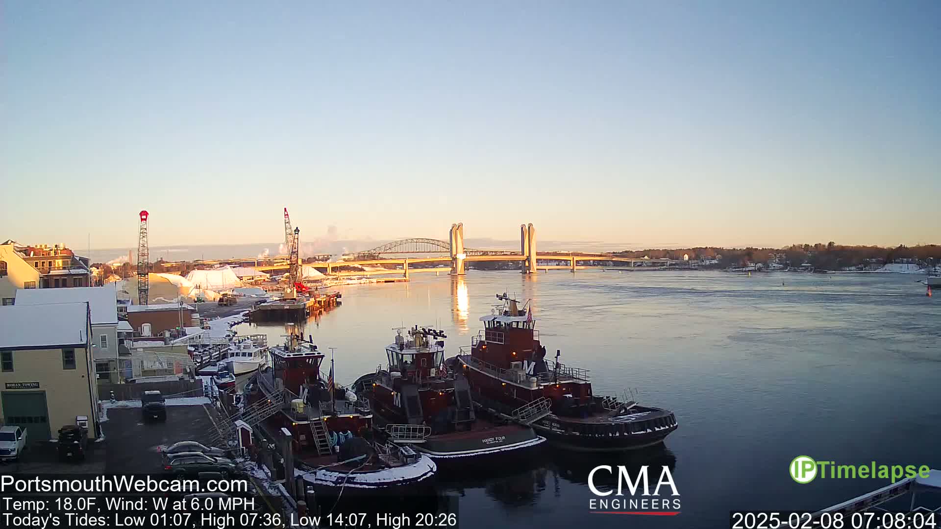 Several tugboats are moored at a dock near a bridge on a calm, cold, and clear morning.