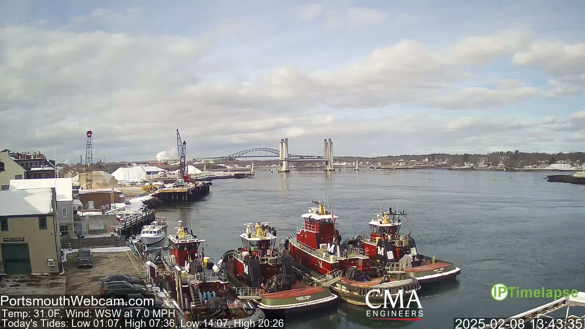Several tugboats are moored at a dock next to a bridge on a partly cloudy day with a temperature of 31 degrees Fahrenheit and a wind blowing from the west-southwest at 7 miles per hour.