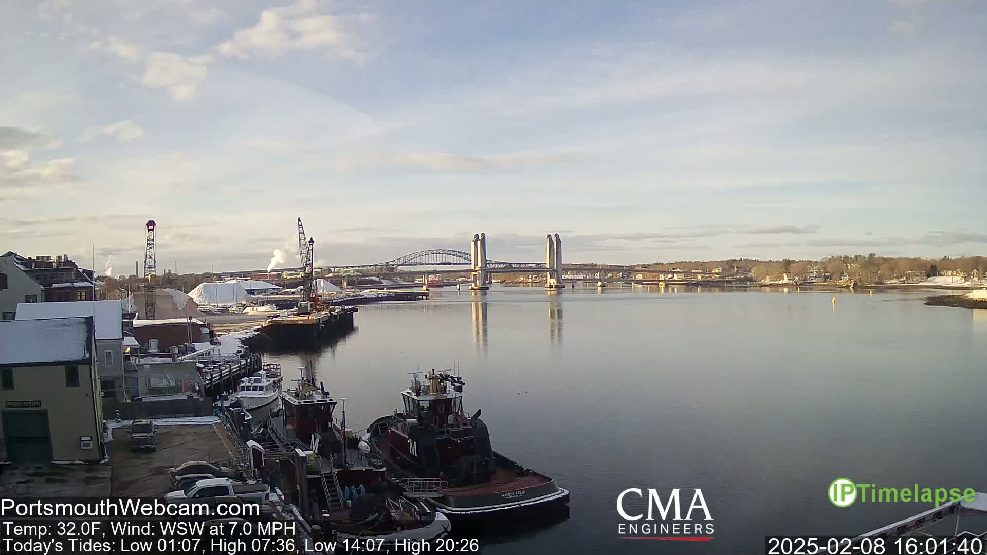 A calm river scene under a partly cloudy sky shows two bridges in the distance, several tugboats docked at a waterfront, and snow-covered buildings and ground.