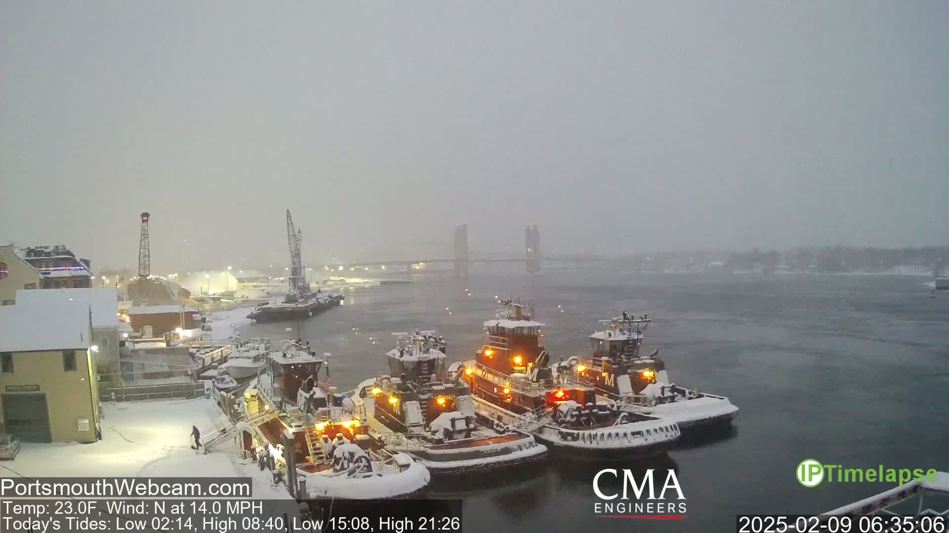Several tugboats are docked in a snow-covered harbor, with a bridge visible in the distance under a heavy, snowy sky.