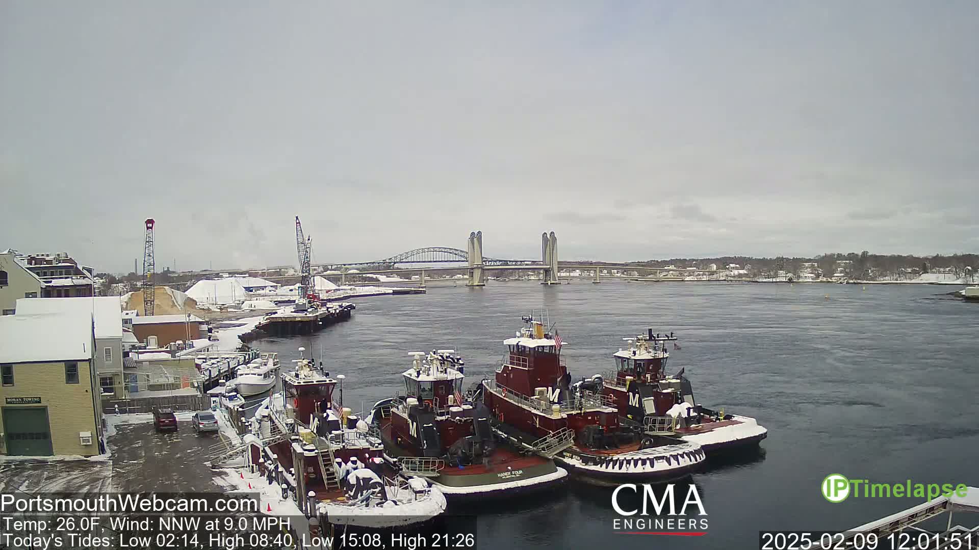 Several tugboats are docked at a snow-covered wharf, with a double-lift bridge in the background under a cloudy sky.
