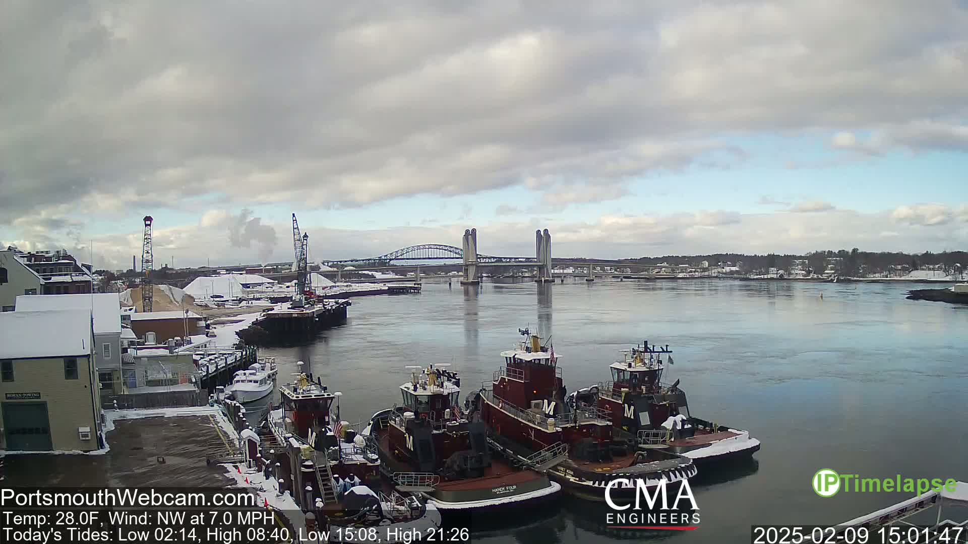 Several tugboats are docked at a snow-covered pier next to a river, under a cloudy sky, with a lift bridge in the background.