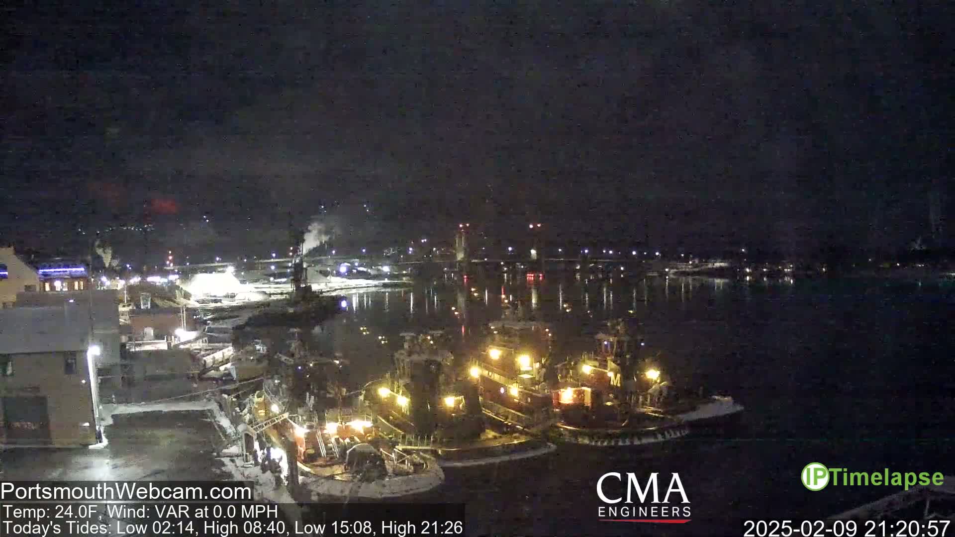 Several tugboats are moored at a dock at night under a dark sky, with a bridge and city lights visible in the distance.
