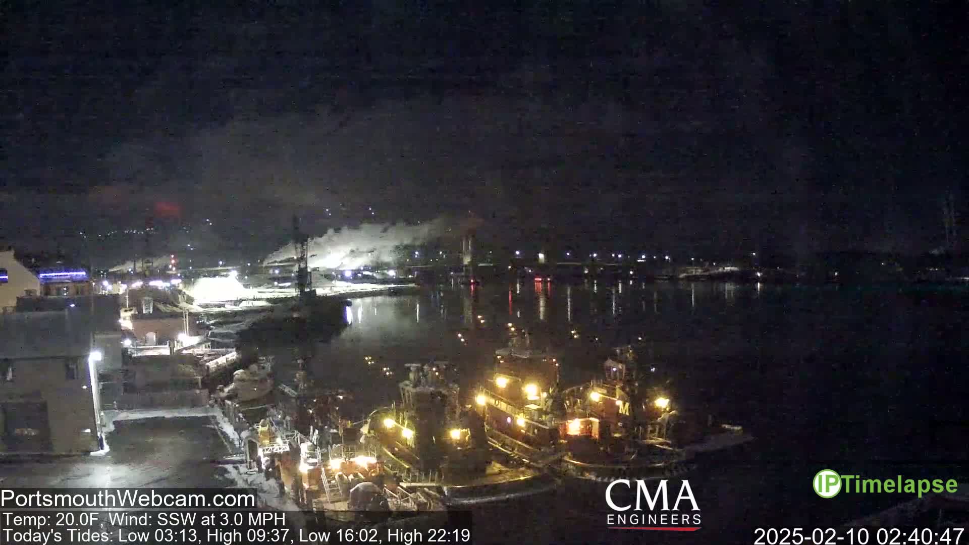 Several tugboats are moored at a dock at night in a calm body of water, with industrial buildings and faint smoke visible in the distance under a dark sky.