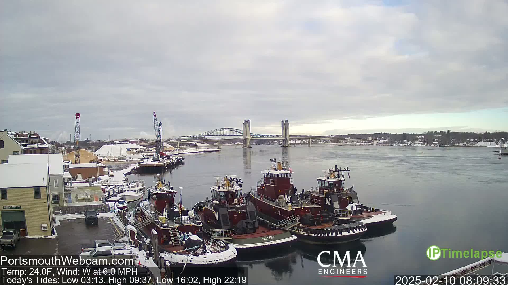 Several tugboats are docked in a snow-covered harbor under an overcast sky, with a bridge visible in the background.