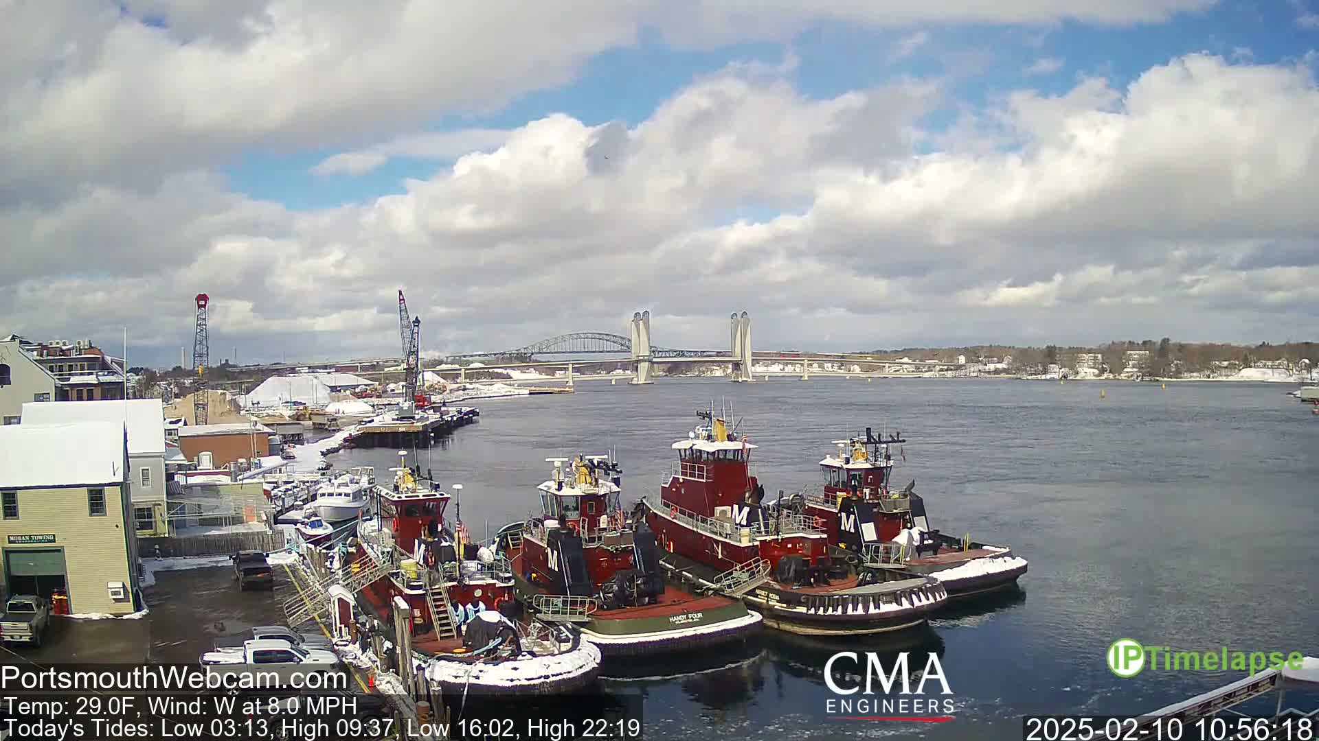 Several tugboats are docked at a snow-dusted waterfront under a partly cloudy sky, with a bridge visible in the distance.