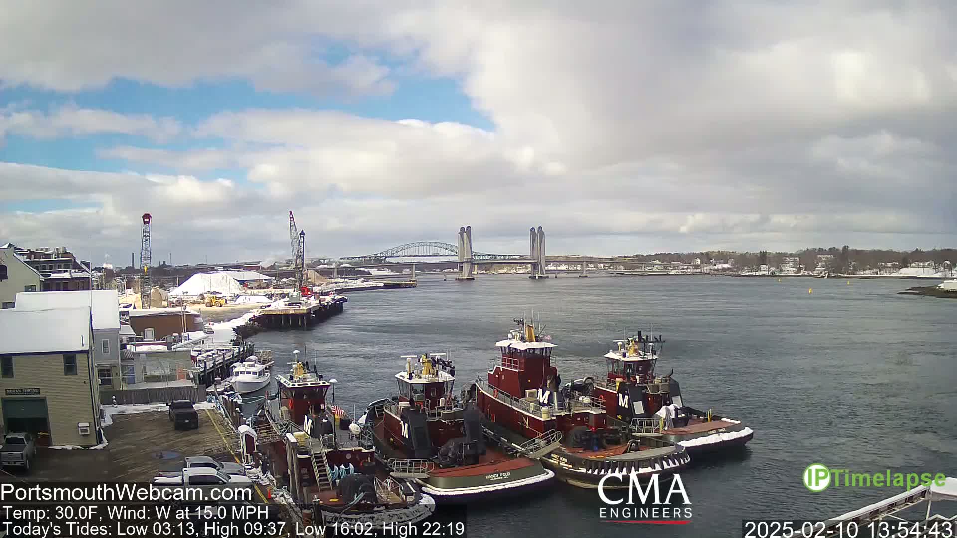 Several tugboats are docked at a waterfront, with a lift bridge spanning a waterway in the background under a partly cloudy sky.
