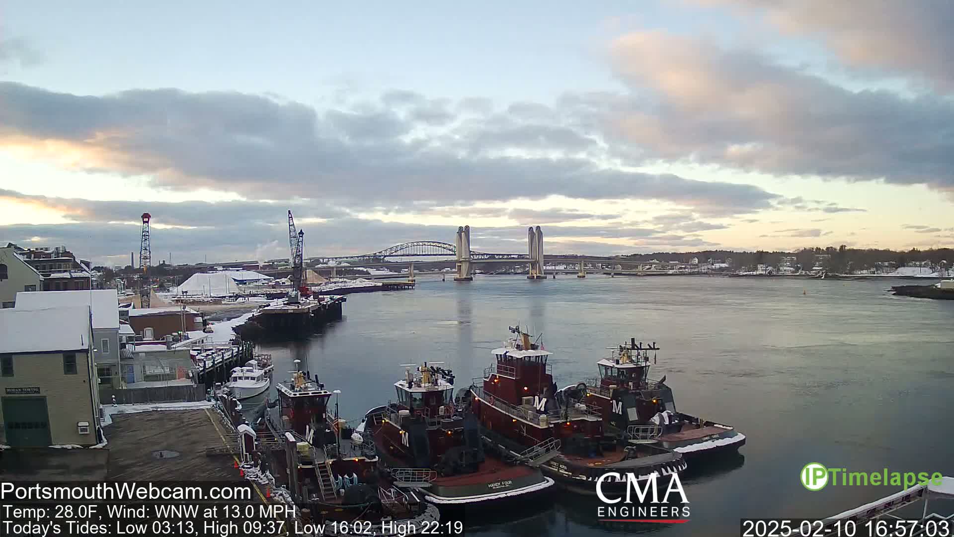 Several tugboats are docked at a snow-dusted waterfront under a partly cloudy sky, with a double-leaf bascule bridge in the background.
