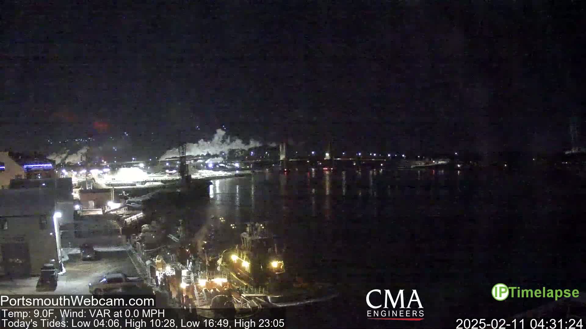 A nighttime view of a harbor with several boats docked, a bridge in the distance, and faint steam rising from industrial areas under a dark sky.