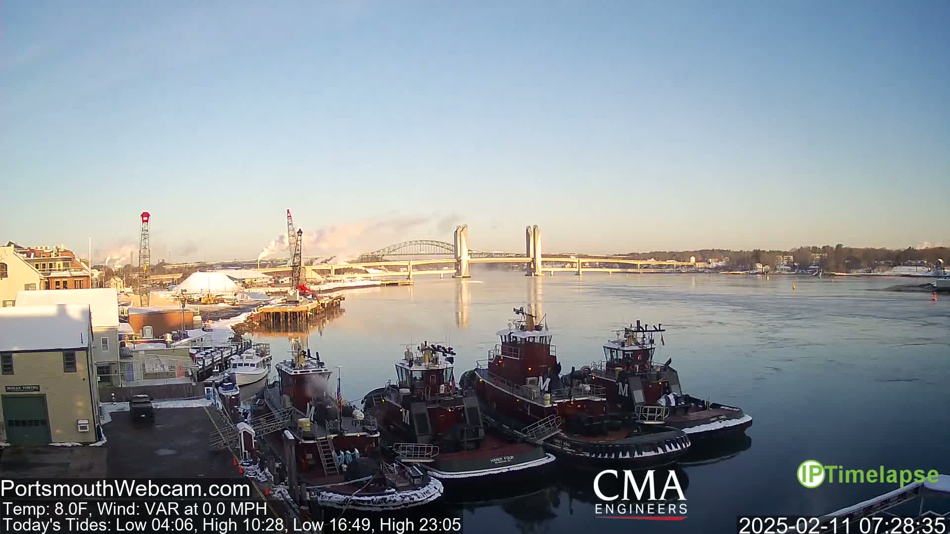 A clear, cold morning shows several tugboats docked near a bridge spanning a wide river, with snow on the ground and some light industrial activity visible in the background.