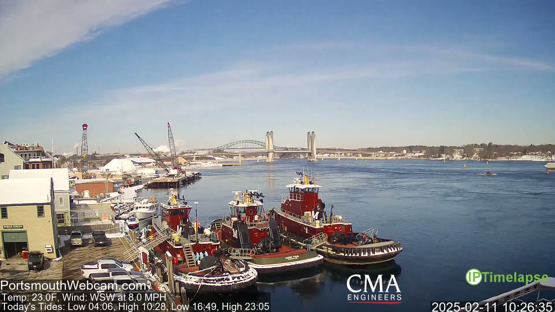 Several red tugboats are docked at a waterfront under a clear blue sky, with a bridge in the distance.
