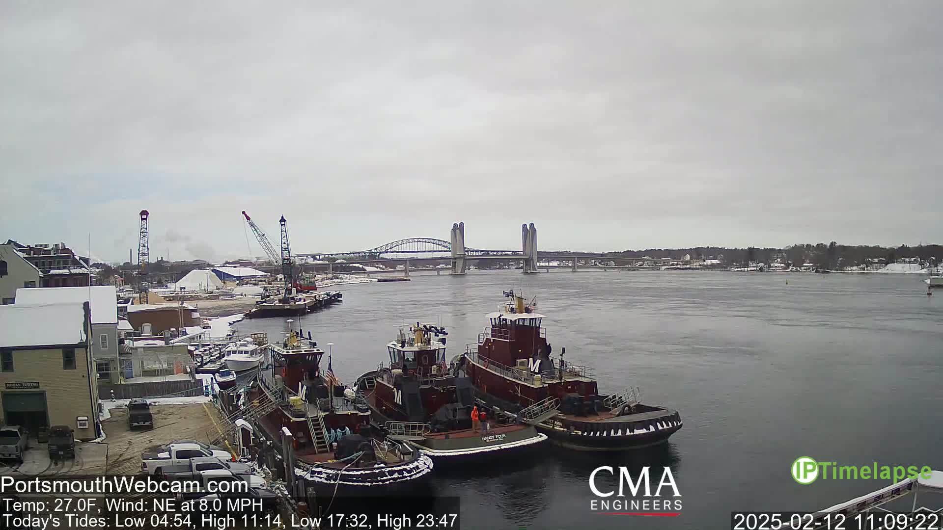 Several tugboats are moored at a dock near a bridge on a cloudy day with a northeast wind.