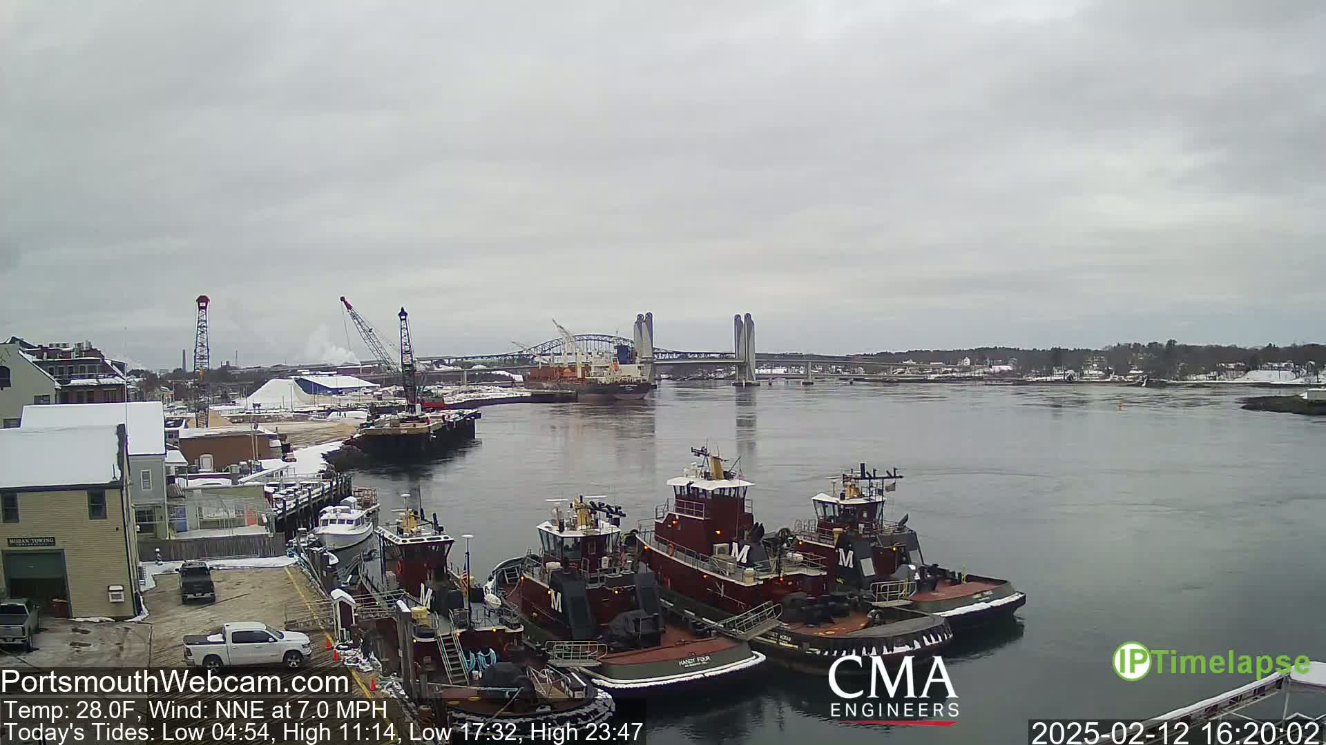 Several tugboats are moored at a dock beside a waterway under an overcast sky, with a bridge and a larger vessel visible in the distance.