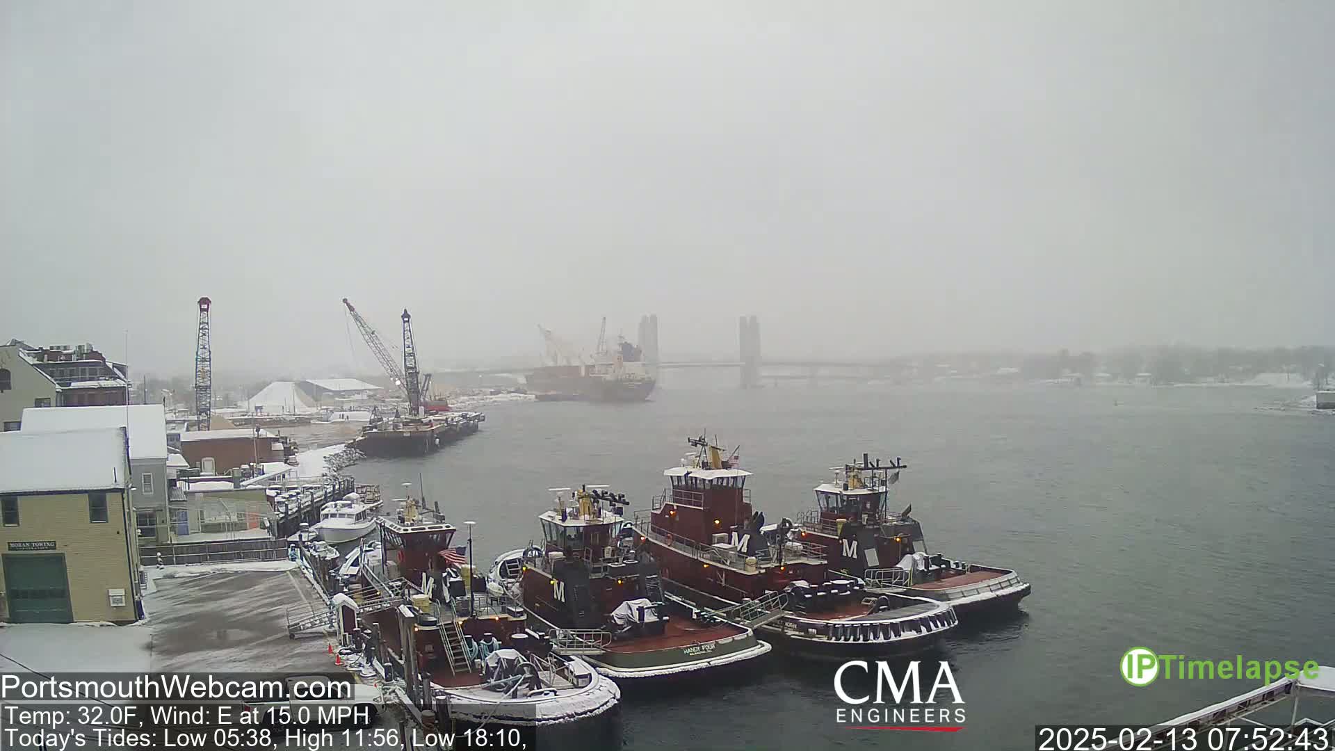 A snow-covered harbor scene shows several tugboats docked, with a larger vessel and a bridge visible in the hazy, snowy distance.