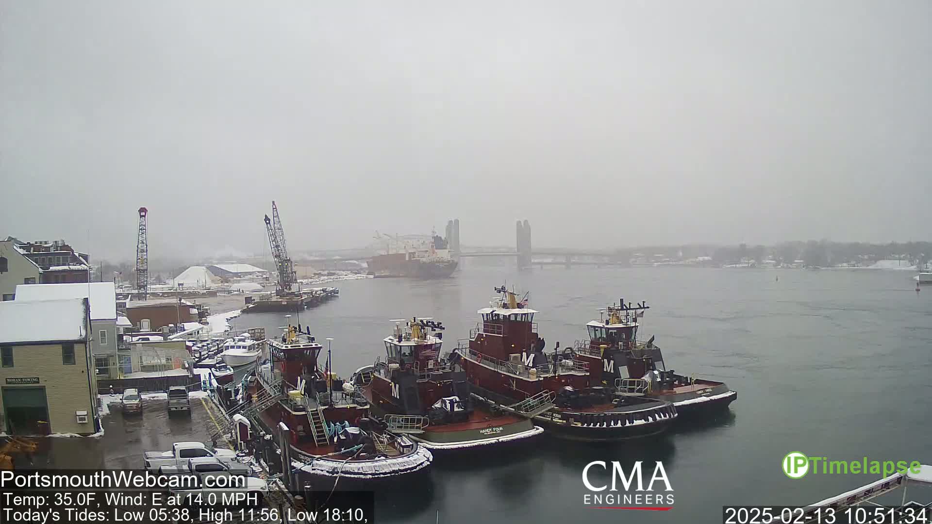 Several tugboats are moored at a snow-covered dock in a harbor, under a gray, snowy sky.