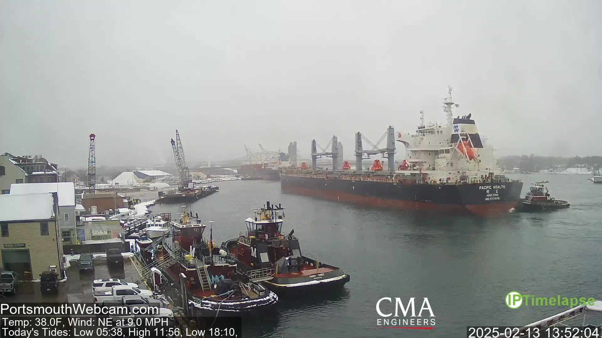 A large cargo ship is being assisted by tugboats in a harbor during snowy conditions.