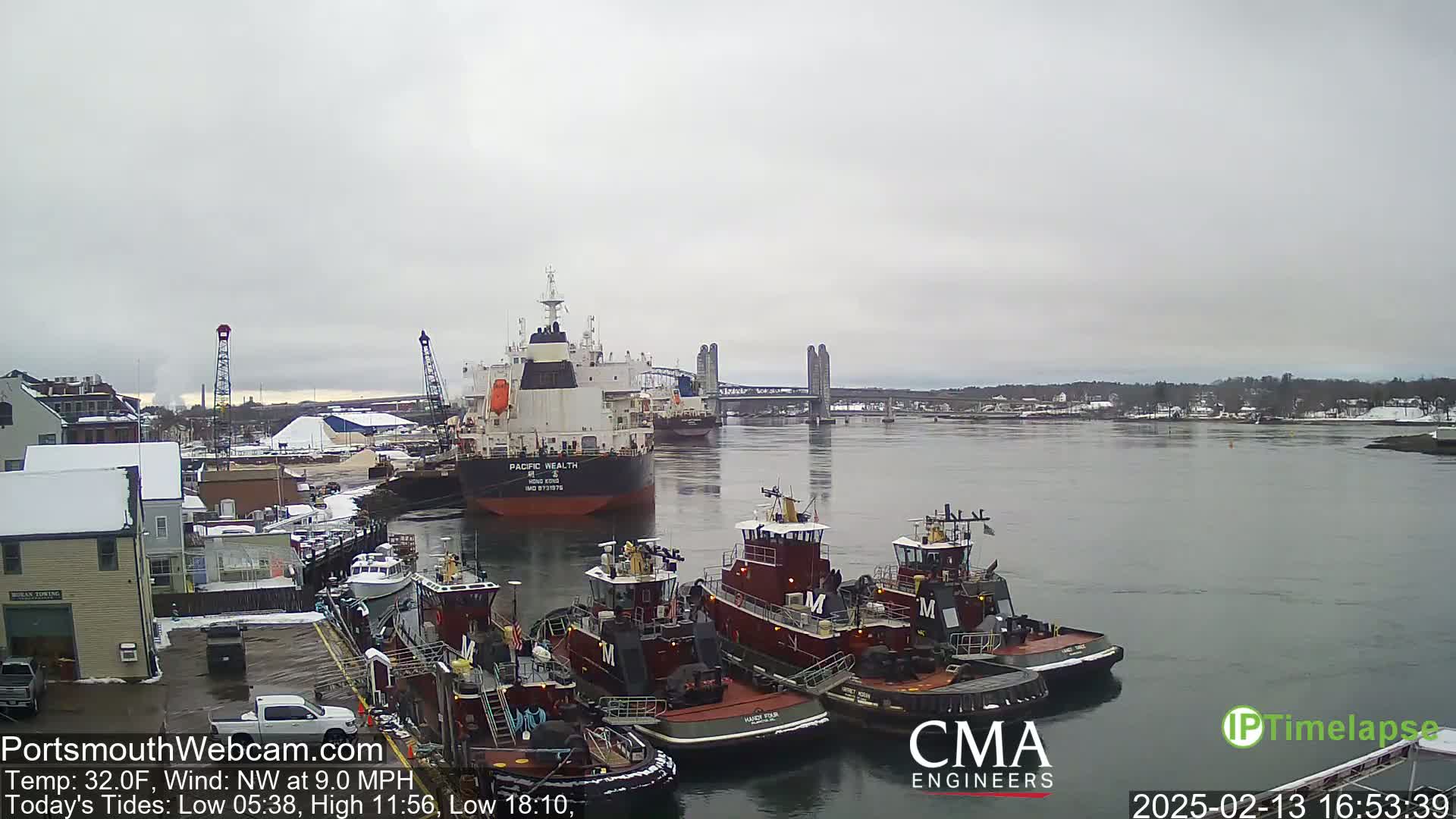 A large cargo ship and several tugboats are moored at a dock in a harbor under an overcast sky, with snow visible on the ground and buildings in the background.