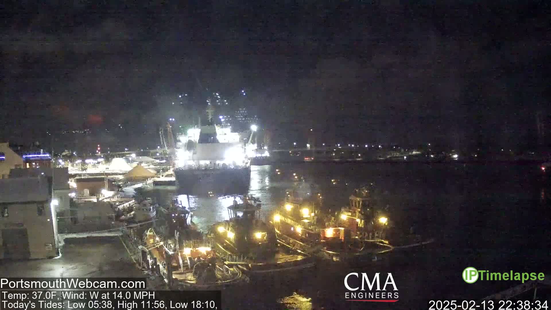 At night, a large cargo ship is docked at a harbor, with several smaller tugboats nearby in calm waters.