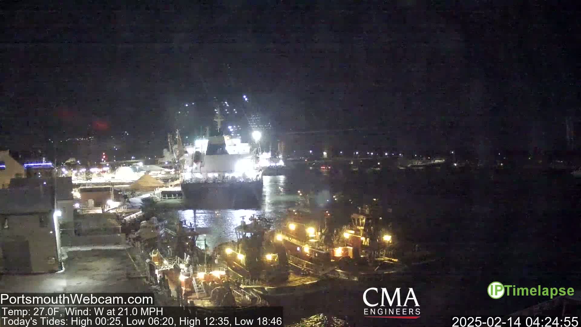 At night, under a dark sky with a 27°F temperature and a 21 mph west wind, several ships, including a large vessel and several tugboats, are illuminated in a harbor.