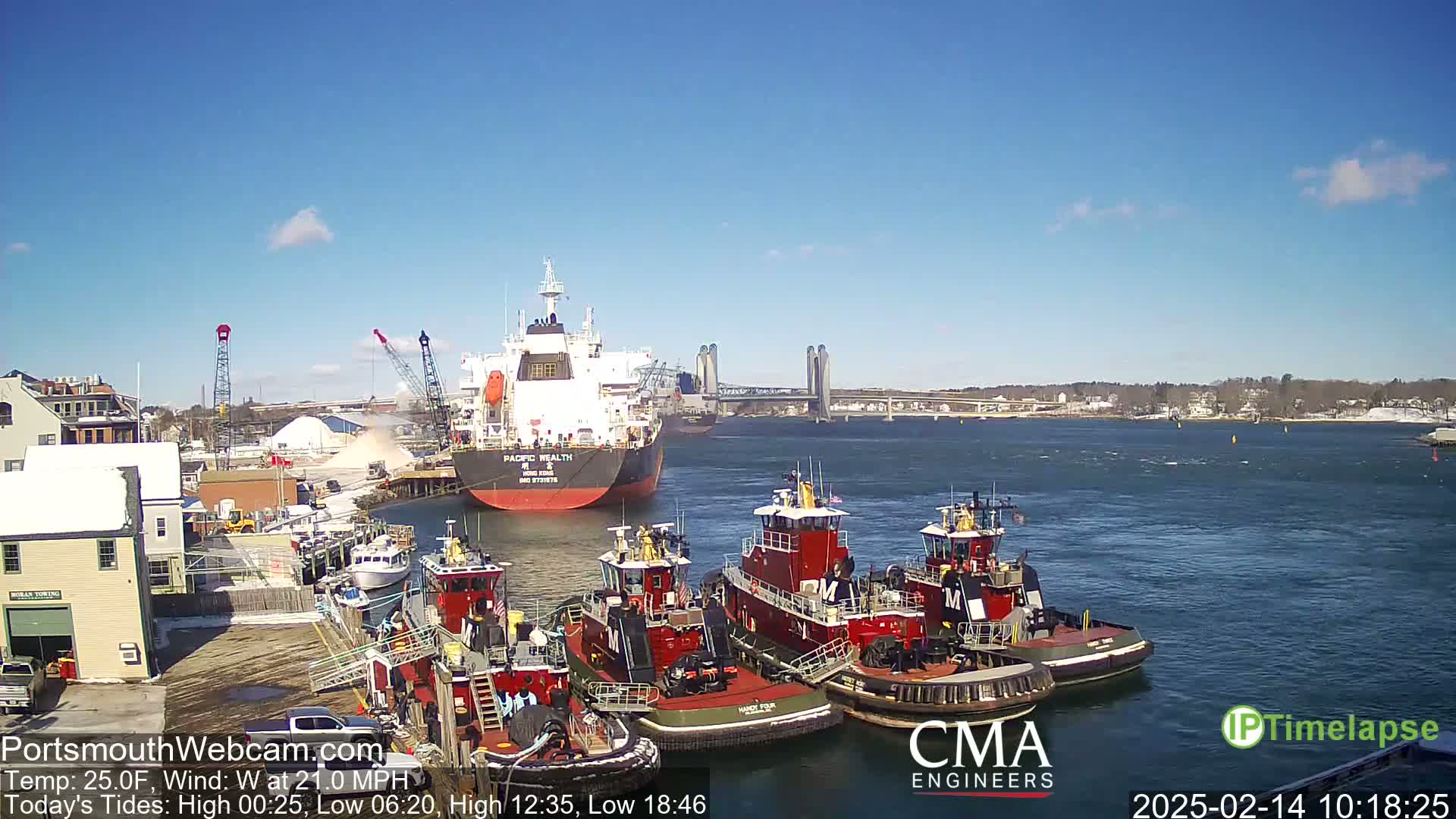 A large cargo ship is docked near several tugboats in a harbor on a sunny day with a light wind.