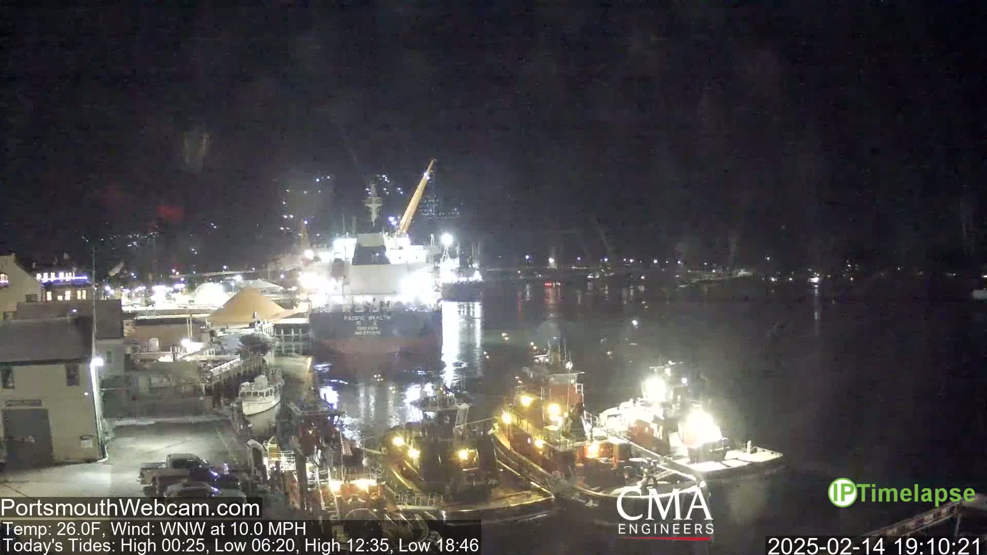 A nighttime harbor scene shows a large cargo ship docked next to smaller tugboats, under a dark, clear sky.