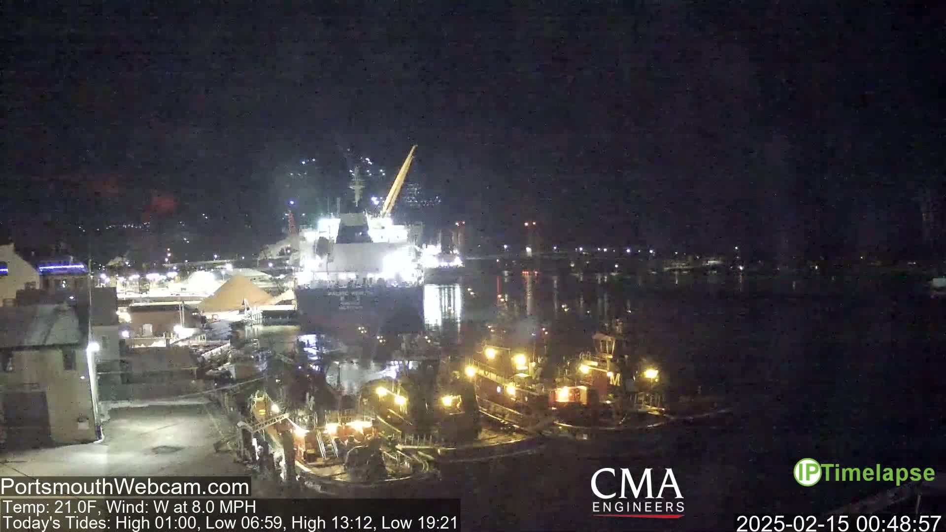 At night, a large cargo ship is docked near several smaller tugboats in a harbor under a clear, dark sky.