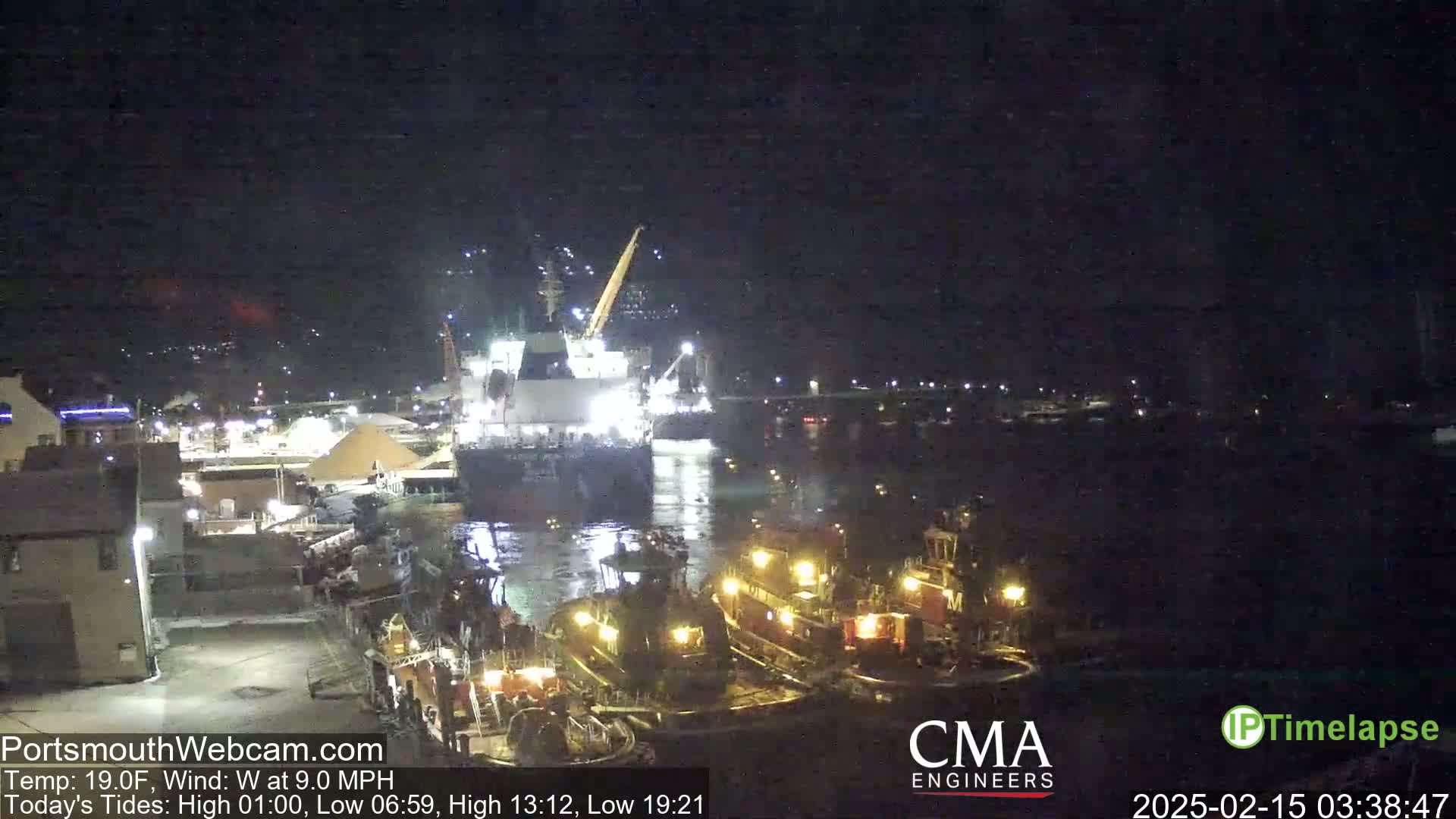 At night, a large cargo ship is docked near several smaller tugboats in a calm harbor.